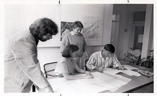 Black and white photo of the founding CDAC director and a student design team working on a project together at a table.